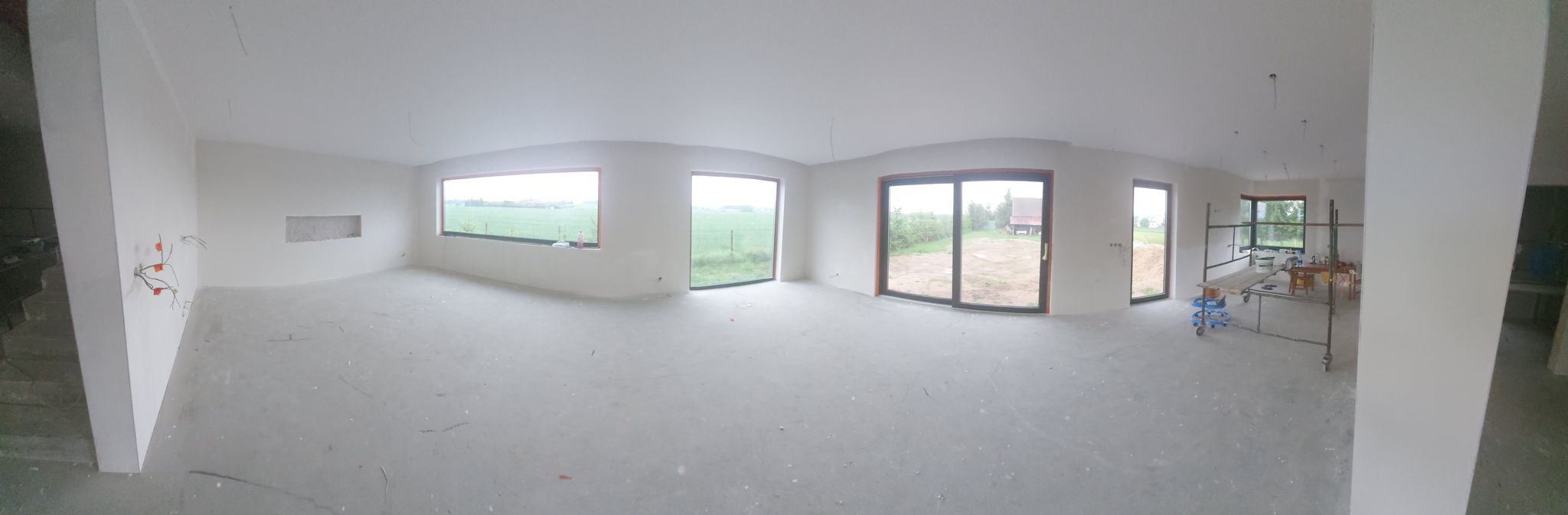 Panoramiczny widok na surowy stan wnętrza domu z widocznymi instalacjami elektrycznymi, oknami z widokiem na zewnątrz i rusztowaniem w tle, przygotowanego do dalszych prac wykończeniowych.