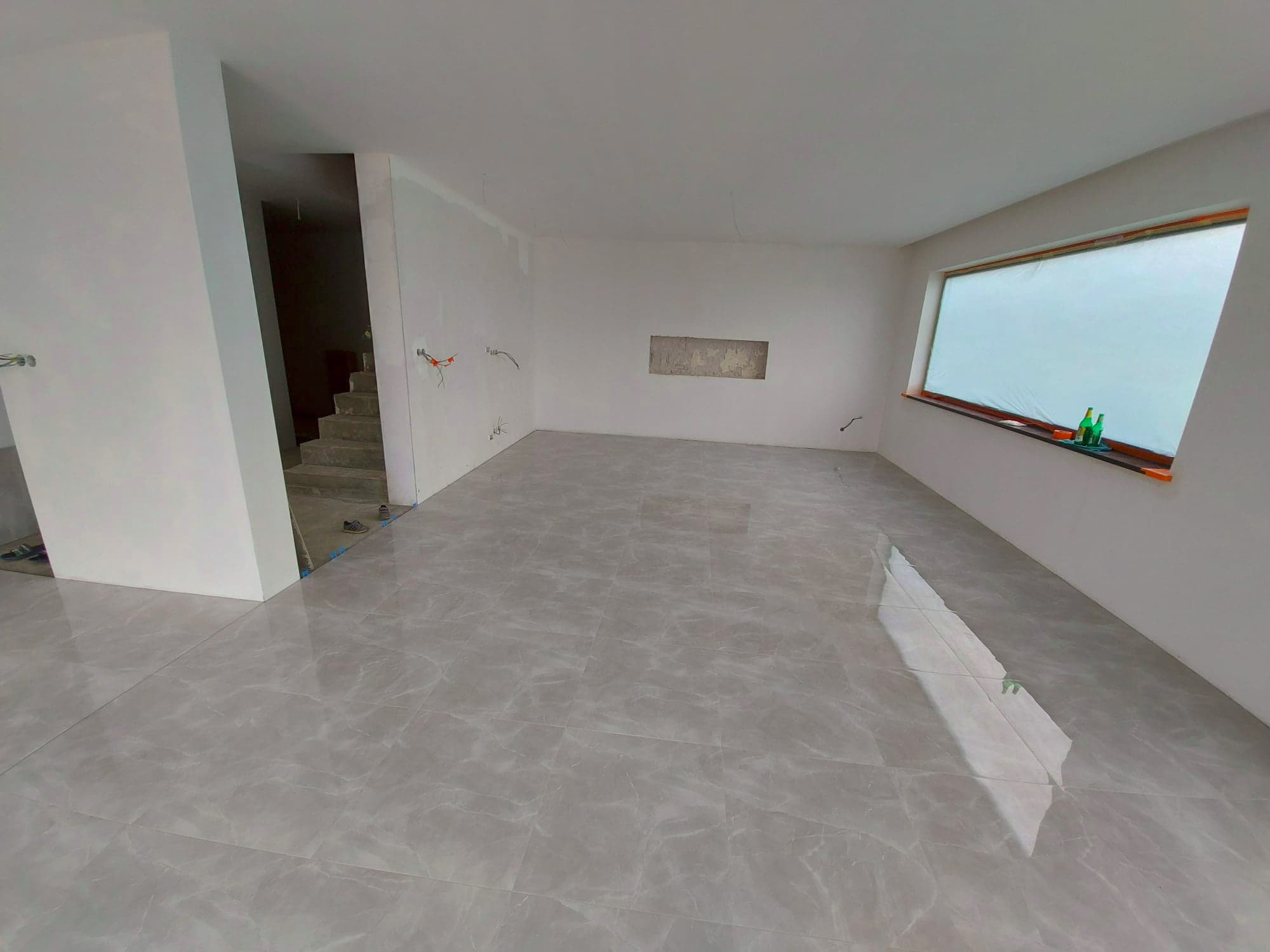 Wykończone płytki podłogowe w jasnoszarym odcieniu w pustym, świeżo wykończonym pomieszczeniu z widocznymi schodami i oknem zabezpieczonym folią.