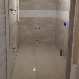 MarMat-BUD - Pusta łazienka w trakcie remontu, wyłożona beżowymi płytkami w dwóch odcieniach, z widocznymi elementami instalacji, zabezpieczona niebieską taśmą malarską przy suficie i z pozostawionymi...