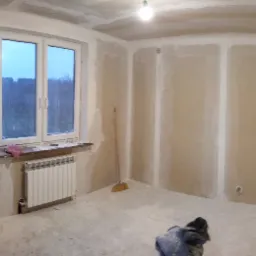 Pokój w trakcie prac wykończeniowych: ściany pokryte płytami gipsowo-kartonowymi, widoczne łączenia wymagające szpachlowania, grzejnik pod oknem, na podłodze ubrania robocze, pomieszczenie...