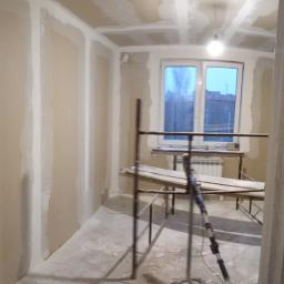 MarMat-BUD - Wnętrze pokoju w trakcie prac wykończeniowych, ściany i sufit pokryte płytami gipsowo-kartonowymi, widoczne szpachlowanie łączeń, metalowe rusztowanie i szlifierka do gipsu, okno z widokiem...