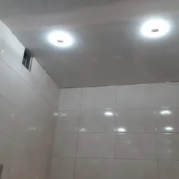 Jasna łazienka z białymi, kwadratowymi płytkami na ścianach, podświetleniem LED nad dekoracyjnymi płytkami z motywem fal na dole, oraz trzema okrągłymi lampami sufitowymi