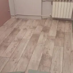 Podłoga wyłożona panelami w odcieniach szarości, widoczny fragment ściany pomalowanej na różowo oraz grzejnik na ścianie w tle, przy ścianie leży młotek.