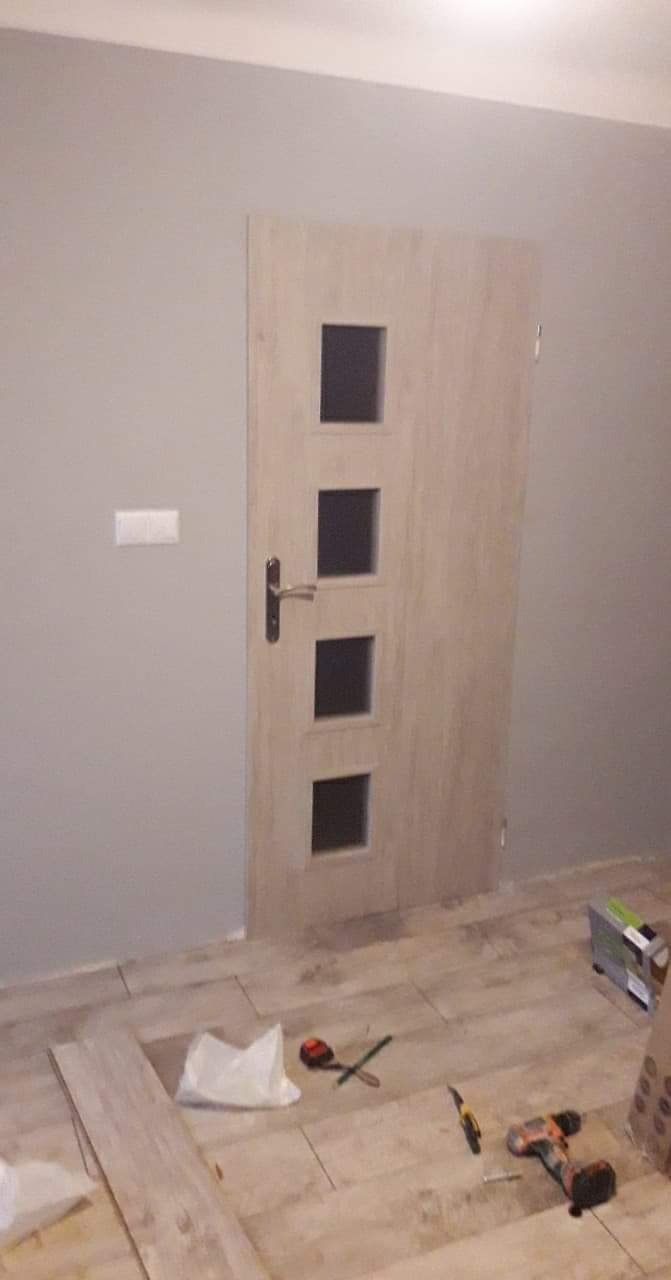 Jasne drzwi wewnętrzne z czterema kwadratowymi przeszkleniami, zamontowane w szarej ścianie, z narzędziami i fragmentami paneli podłogowych na podłodze.