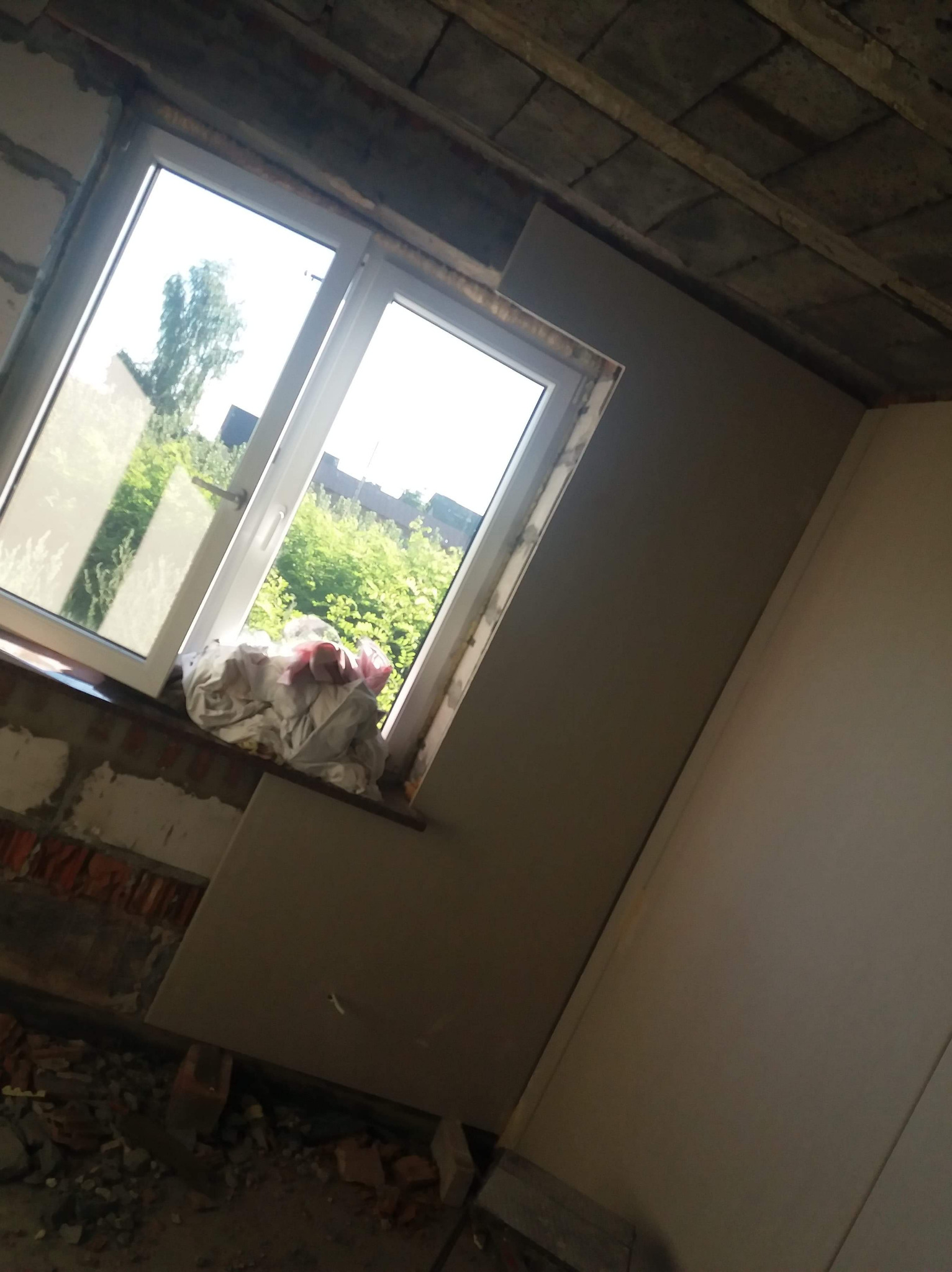 Wnętrze w trakcie prac wykończeniowych: ściana częściowo pokryta płytą kartonowo-gipsową, widoczne surowe cegły i pustaki, okno z widokiem na zieleń, gruz na podłodze.