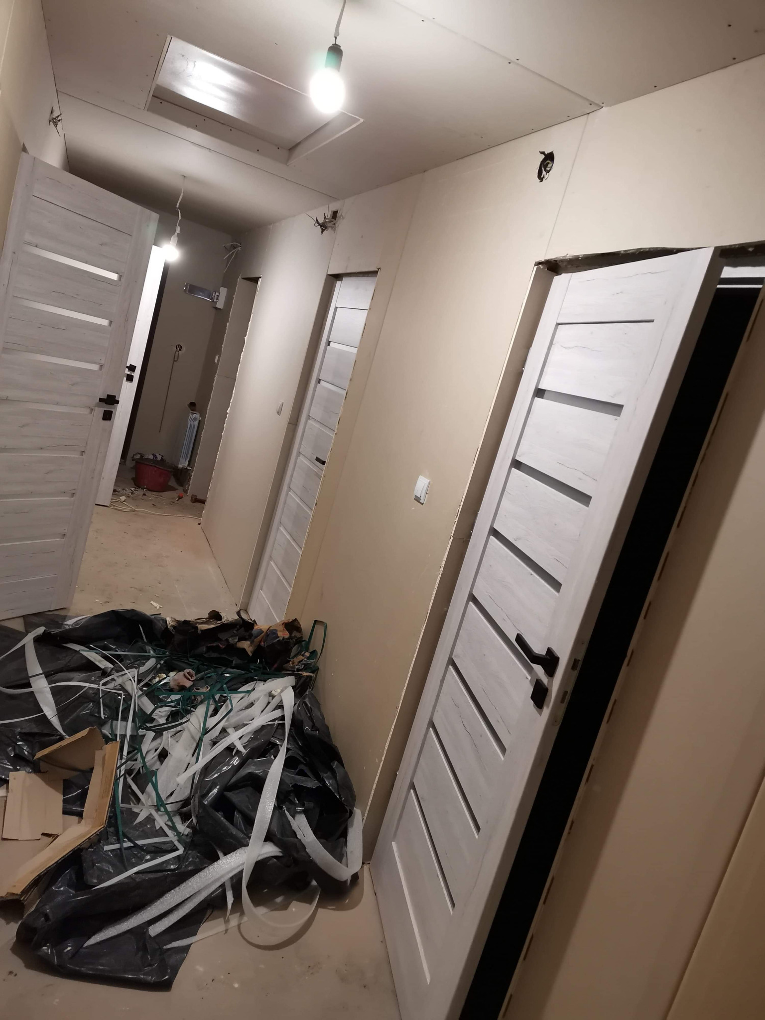 Korytarz w trakcie remontu z zamontowanymi białymi drzwiami z poziomymi frezami, widoczne ślady prac wykończeniowych i pozostałości po folii zabezpieczającej.