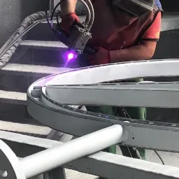 Pracownik w kombinezonie ochronnym i masce spawalniczej podczas obróbki metalowego elementu o kształcie okręgu, z widocznym intensywnym fioletowym światłem procesu i iskrami.