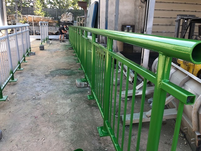 Malowanie proszkowe zielonej metalowej balustrady na zewnątrz, widoczny pracownik w tle podczas aplikacji farby.