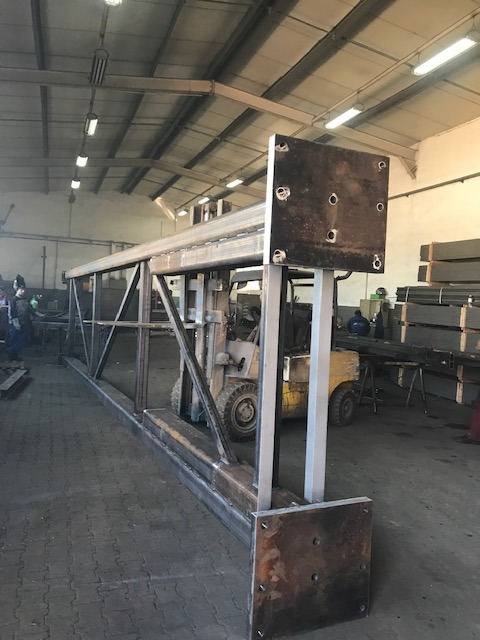 Konstrukcja stalowa kratowa z kwadratowymi płytami mocującymi, widoczna w hali produkcyjnej z wózkiem widłowym w tle.