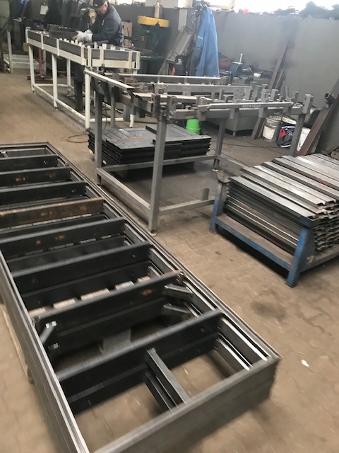 Warsztatowa sceneria z pracownikiem w tle, obróbka metalu: spawanie profili do konstrukcji bramy, widoczne elementy ramy bramy, profile stalowe na regale i stanowisko pracy.