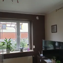 Pokój po remoncie z brązowymi ścianami, widoczne okno z roślinami na parapecie, drabina malarska oparta o ścianę, ślady po taśmie malarskiej na ścianie.