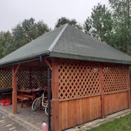 Drewniana altana z zielonym dachem, kratowanymi ścianami i wyposażeniem w postaci stołu, ławek, grilla i skrzynki piwa Perła, na wybrukowanym patio.