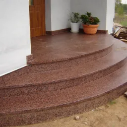 Zaokrąglone, trzybiegowe schody kamienne z czerwonego granitu prowadzące do wejścia domu w Białymstoku, z doniczkami na górnym podeście. W tle widoczne deski i fragment otoczenia.