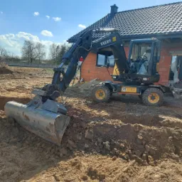 Czarna koparka Volvo na tle ceglanego domu w trakcie budowy, łyżka zagłębiona w ziemi, widoczny wykop