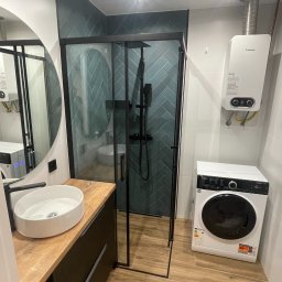Extremo - Nowoczesna łazienka z kabiną prysznicową typu walk-in, umywalką nablatową na szafce z drewnianym blatem, pralką i bojlerem Junkers. Ściany wyłożone płytkami w odcieniach zieleni ułożonymi w jodełkę.