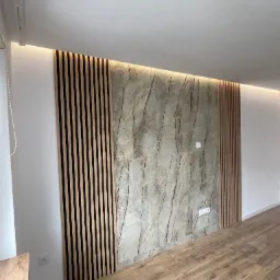Ściana z dekoracyjną okładziną kamienną i drewnianymi lamelami, podświetlona listwą LED, nad parkietem imitującym drewno. Nowoczesny design z gniazdkami elektrycznymi.