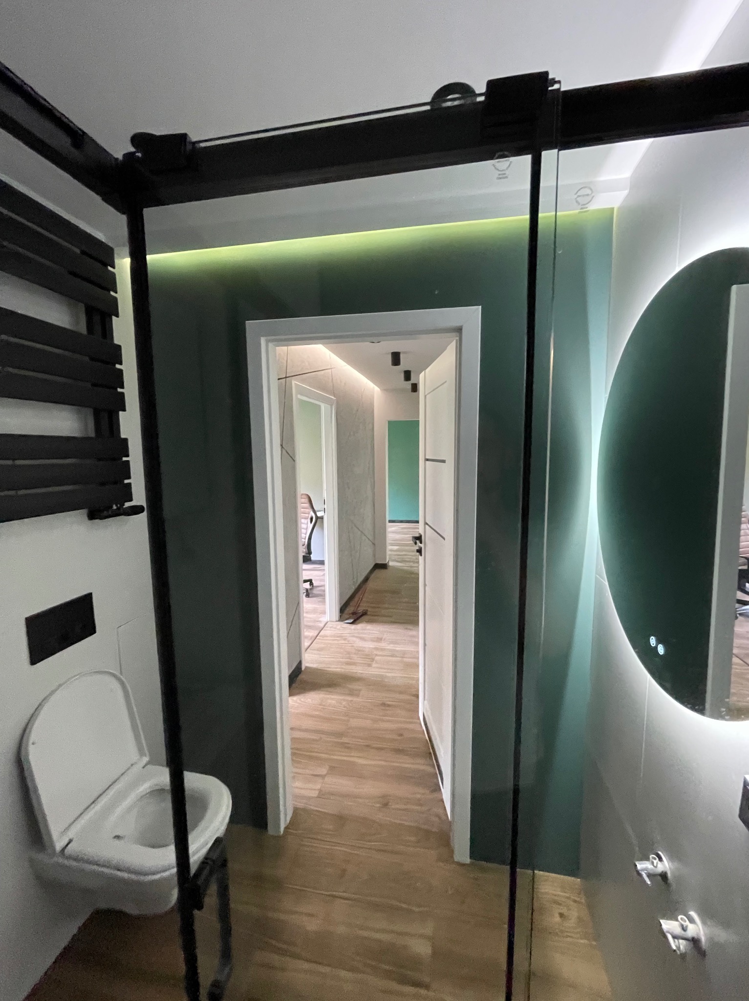 Nowoczesna łazienka z kabiną prysznicową typu walk-in, podświetlanym lustrem i grzejnikiem drabinkowym w kolorze czarnym, z widokiem na dalsze pomieszczenia.