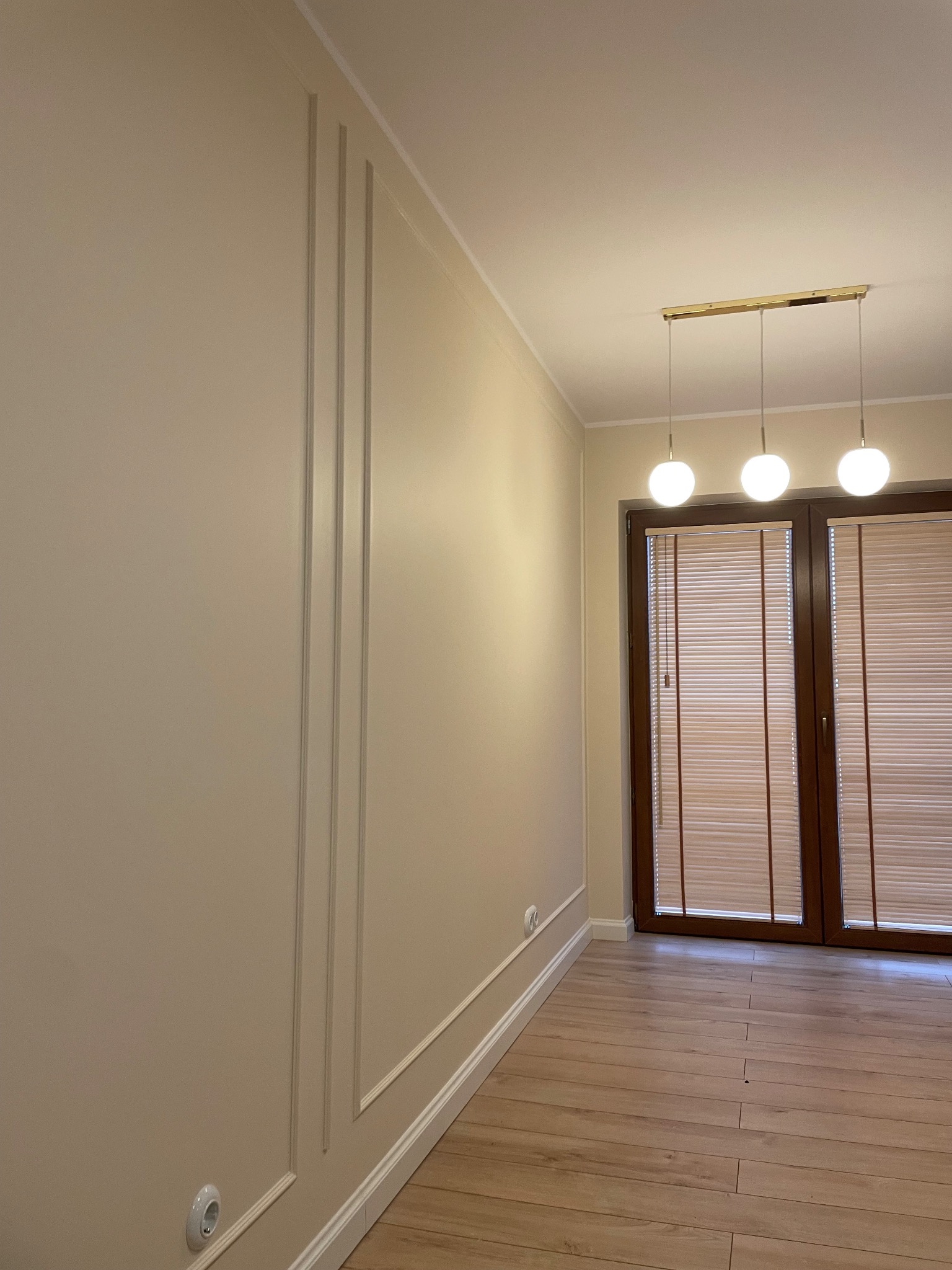 Beżowa ściana z dekoracyjnymi pionowymi listwami, drewniana podłoga i okno z żaluzjami w nowocześnie wykończonym wnętrzu.