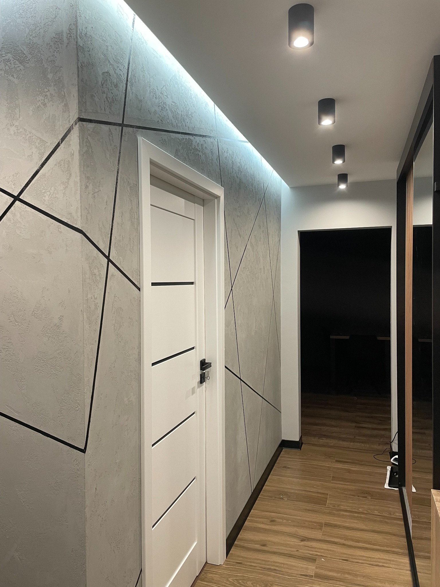 Wnętrze korytarza z dekoracyjną, szarą ścianą z czarnymi liniami, białe drzwi z poziomymi frezowaniami, drewniana podłoga i oświetlenie sufitowe.