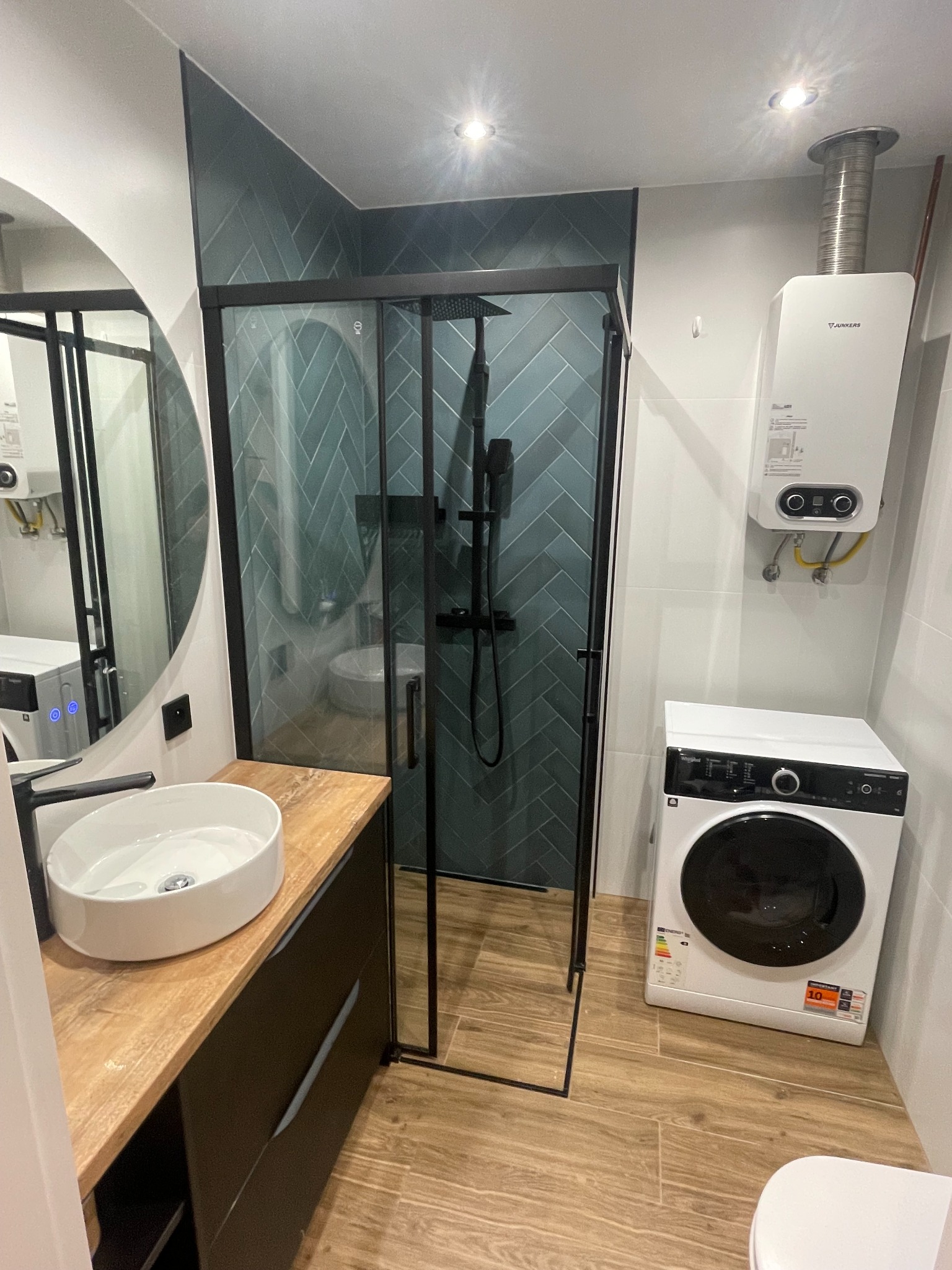 Nowoczesna łazienka z kabiną prysznicową typu walk-in, umywalką nablatową na szafce z drewnianym blatem, pralką i bojlerem Junkers. Ściany wyłożone płytkami w odcieniach zieleni ułożonymi w jodełkę.