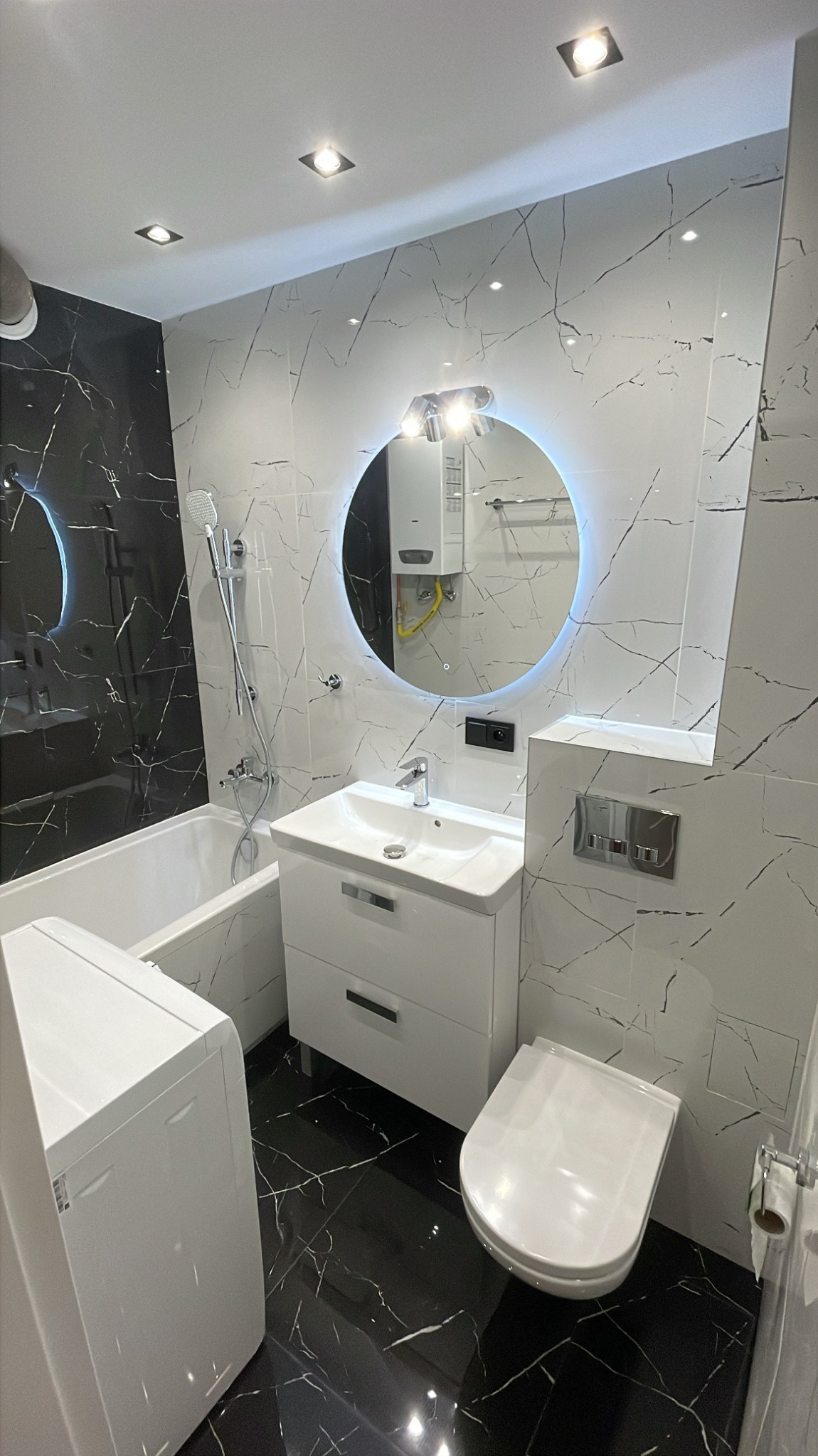 Nowoczesna łazienka z czarno-białymi marmurkowymi płytkami, okrągłym lustrem z podświetleniem LED, białą armaturą i pralką.