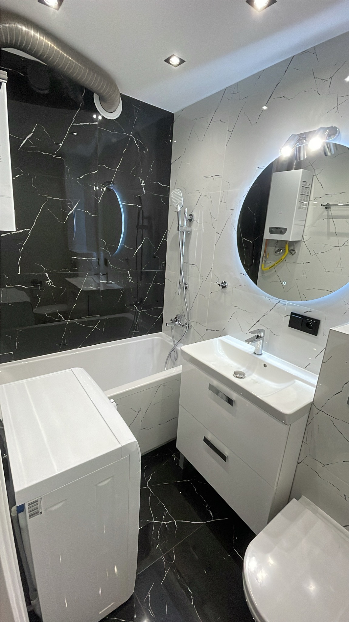 Nowoczesna łazienka z czarno-białymi płytkami imitującymi marmur, okrągłym lustrem z podświetleniem LED, białą umywalką z szafką, wanną i pralką.