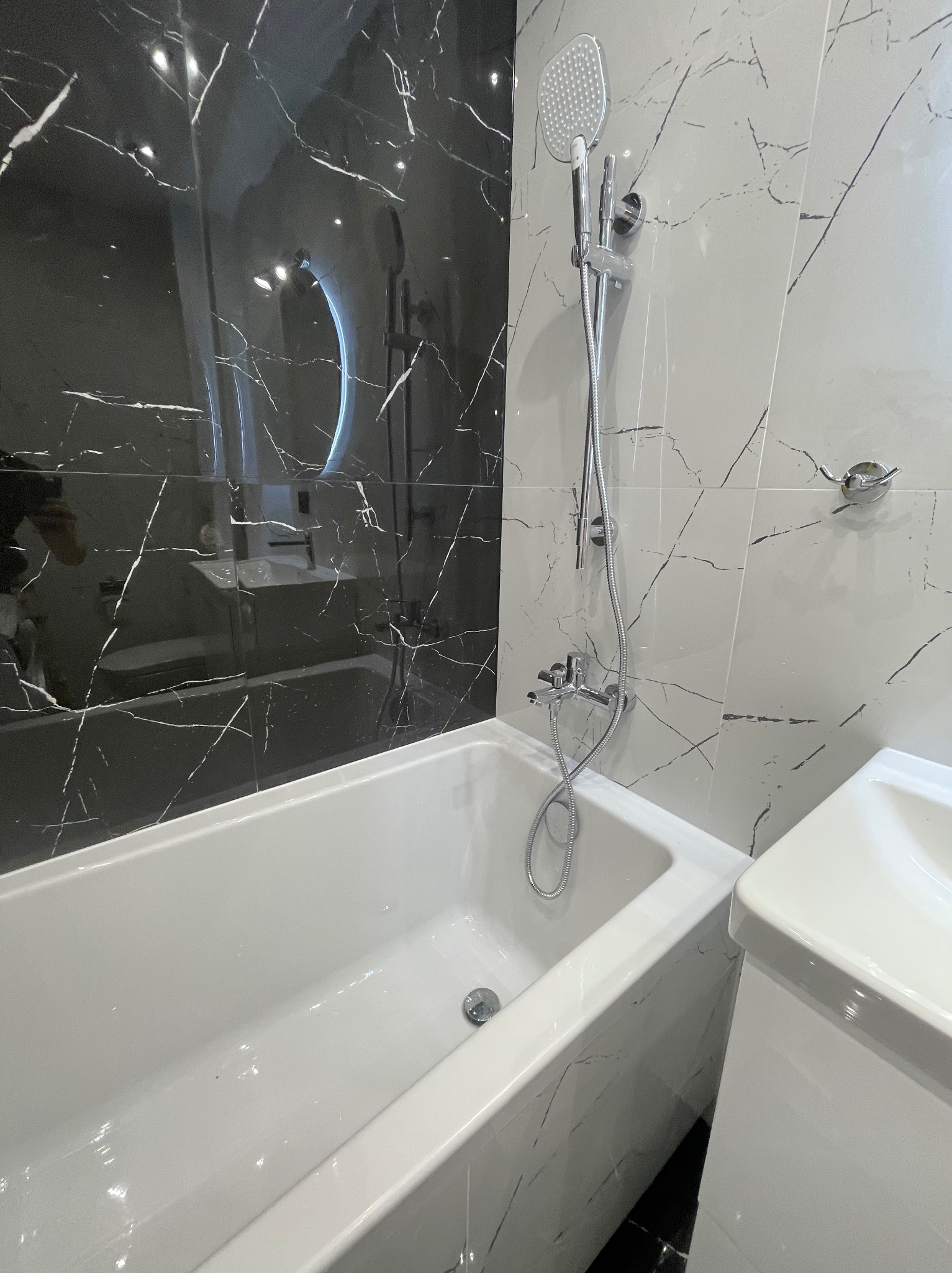 Nowoczesna łazienka z wanną, deszczownicą i umywalką, wykończona czarno-białymi płytkami imitującymi marmur.