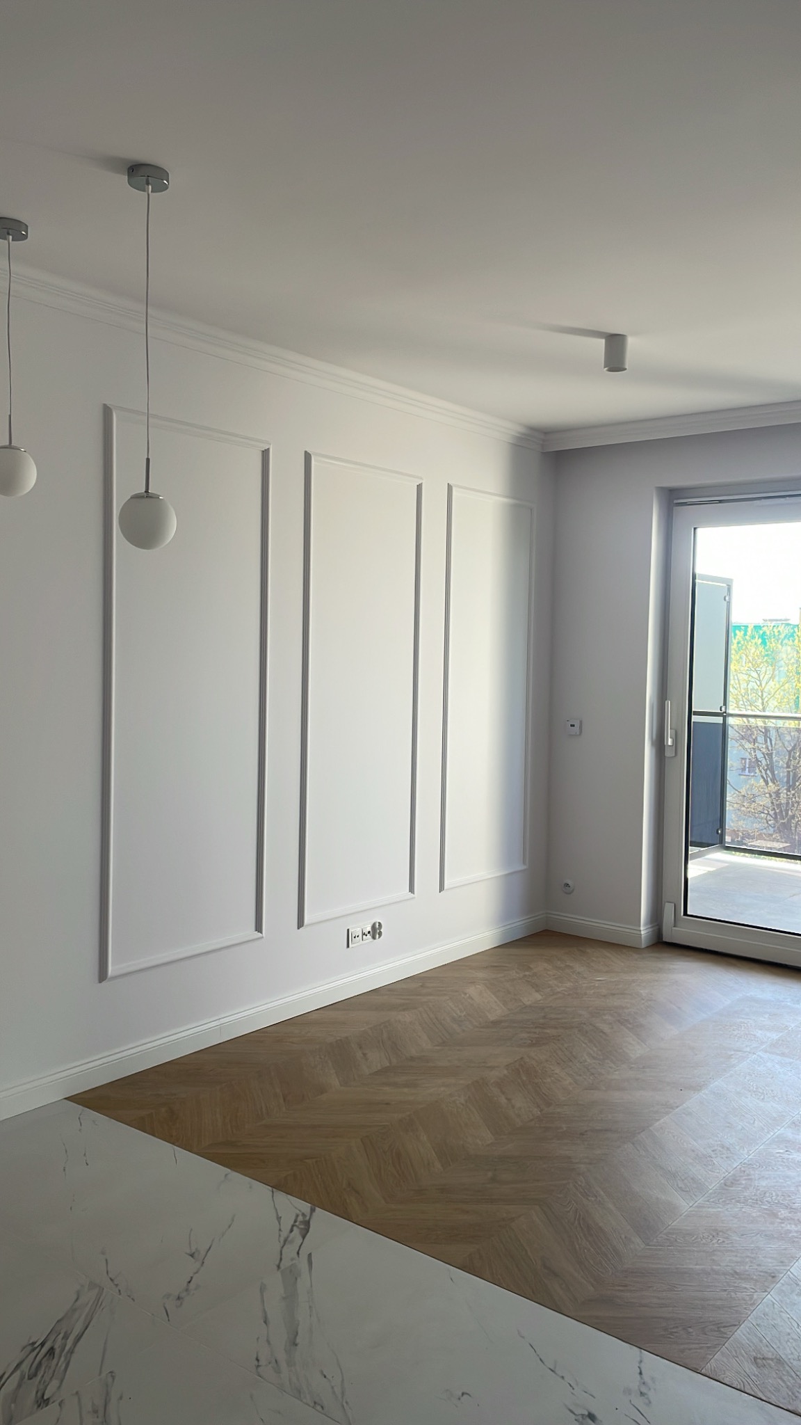 Jasne, minimalistyczne wnętrze z parkietem w jodełkę, ozdobnymi panelami ściennymi i widokiem na balkon. Dominuje biel i naturalne odcienie drewna.