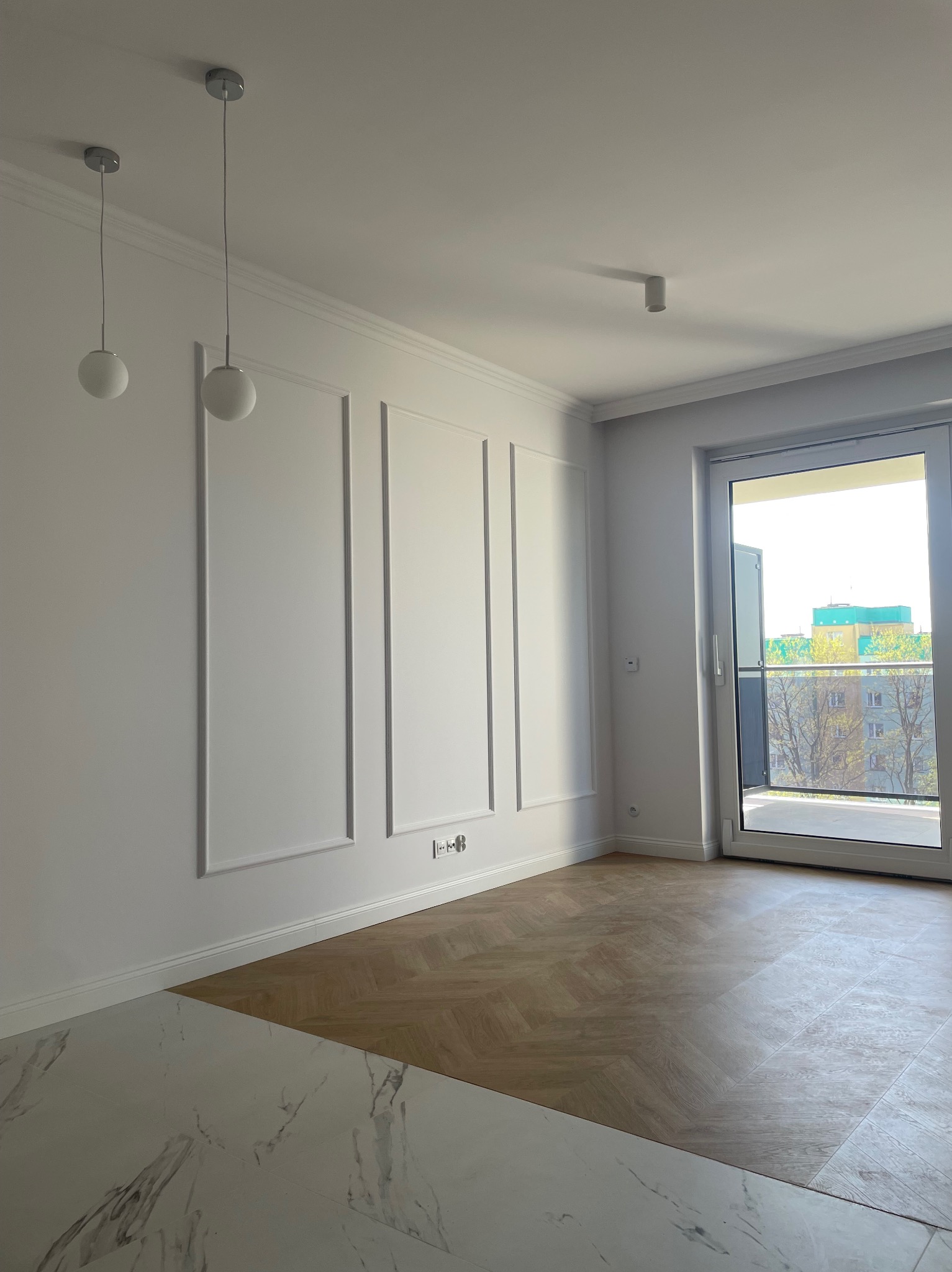 Białe wnętrze po remoncie: klasyczne panele ścienne, parkiet w jodełkę i marmurowe płytki podłogowe. Widok na balkon i budynek za oknem, minimalistyczne lampy.