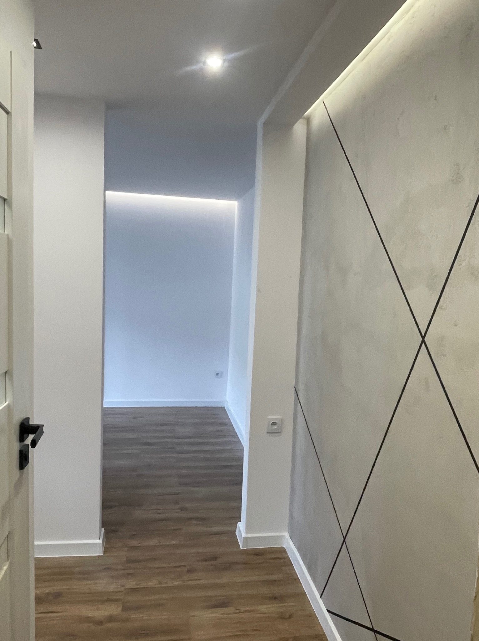 Korytarz po remoncie z drewnianą podłogą i ścianą z geometrycznym wzorem. Widoczne oświetlenie LED i białe listwy przypodłogowe. Częściowo widoczne drzwi.