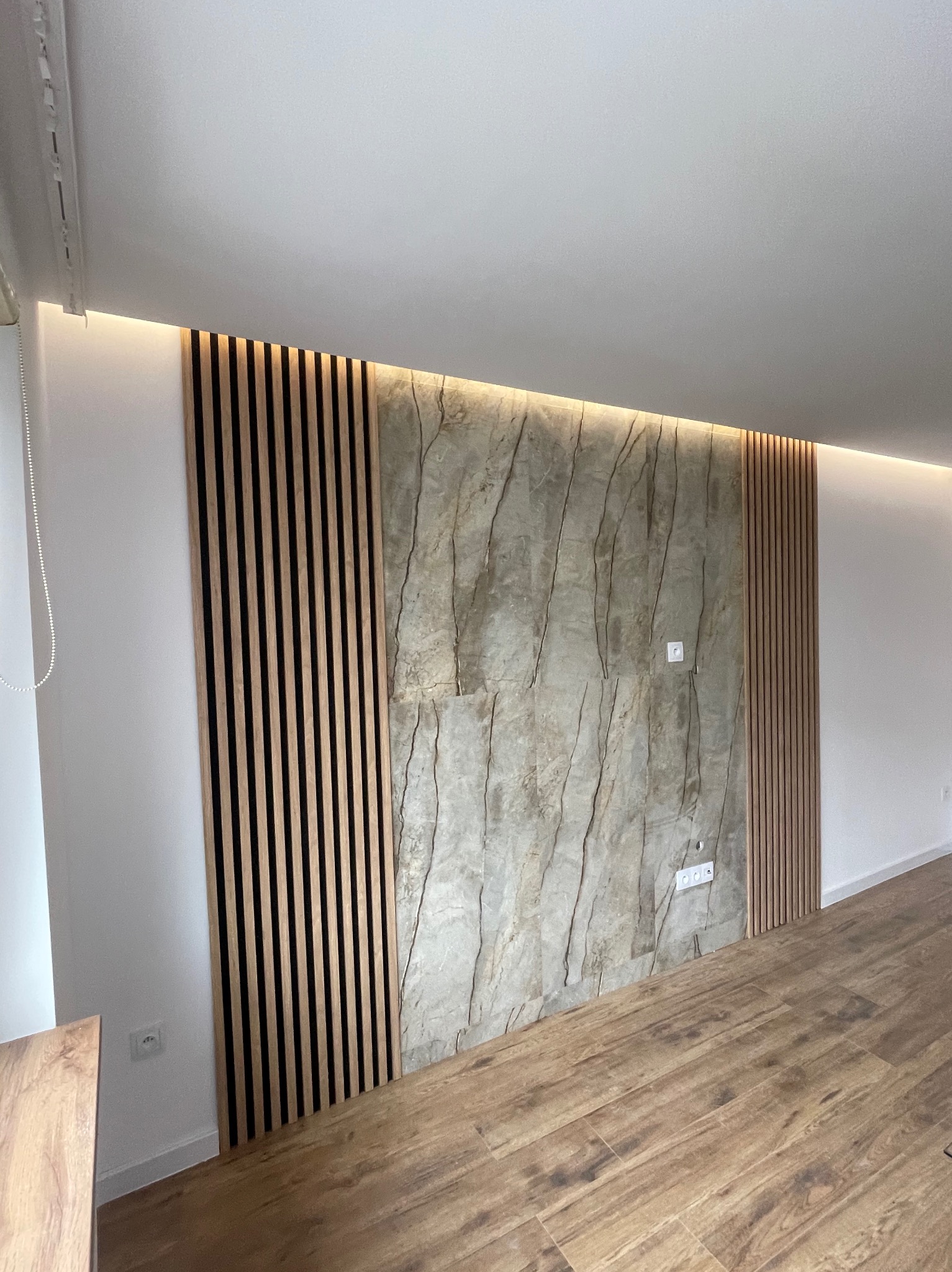 Ściana z dekoracyjną okładziną kamienną i drewnianymi lamelami, podświetlona listwą LED, nad parkietem imitującym drewno. Nowoczesny design z gniazdkami elektrycznymi.