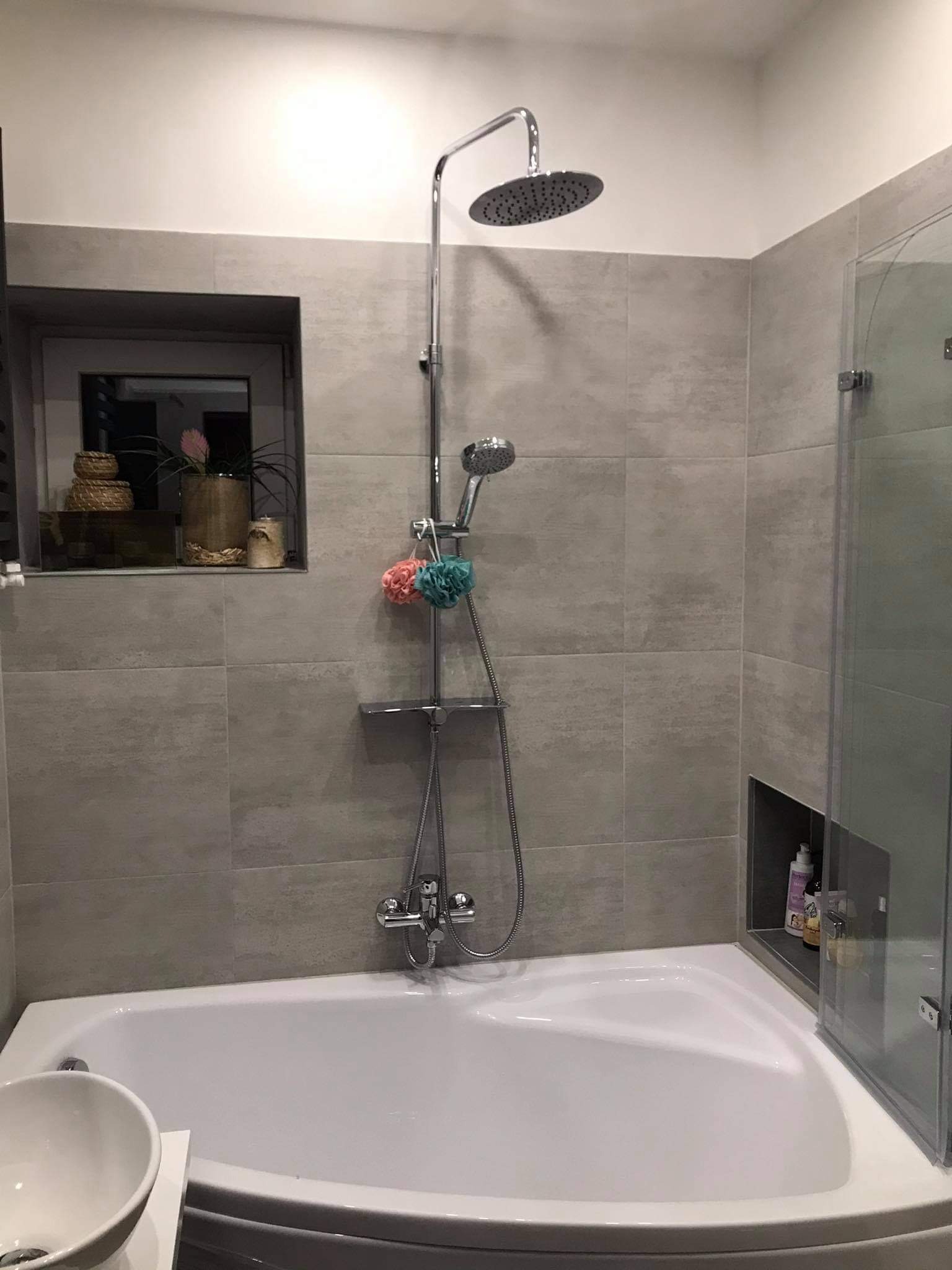 Jasna łazienka z wanną narożną, szarymi płytkami na ścianach, chromowaną armaturą prysznicową i dekoracyjnymi elementami w półce okiennej oraz wnęce prysznicowej.