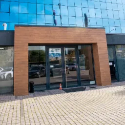 Wejście do biurowca z przeszkloną fasadą, drewnianym obramowaniem wejścia i widocznymi w szybach odbiciami samochodów. Na chodniku stoi pomarańczowy pachołek.