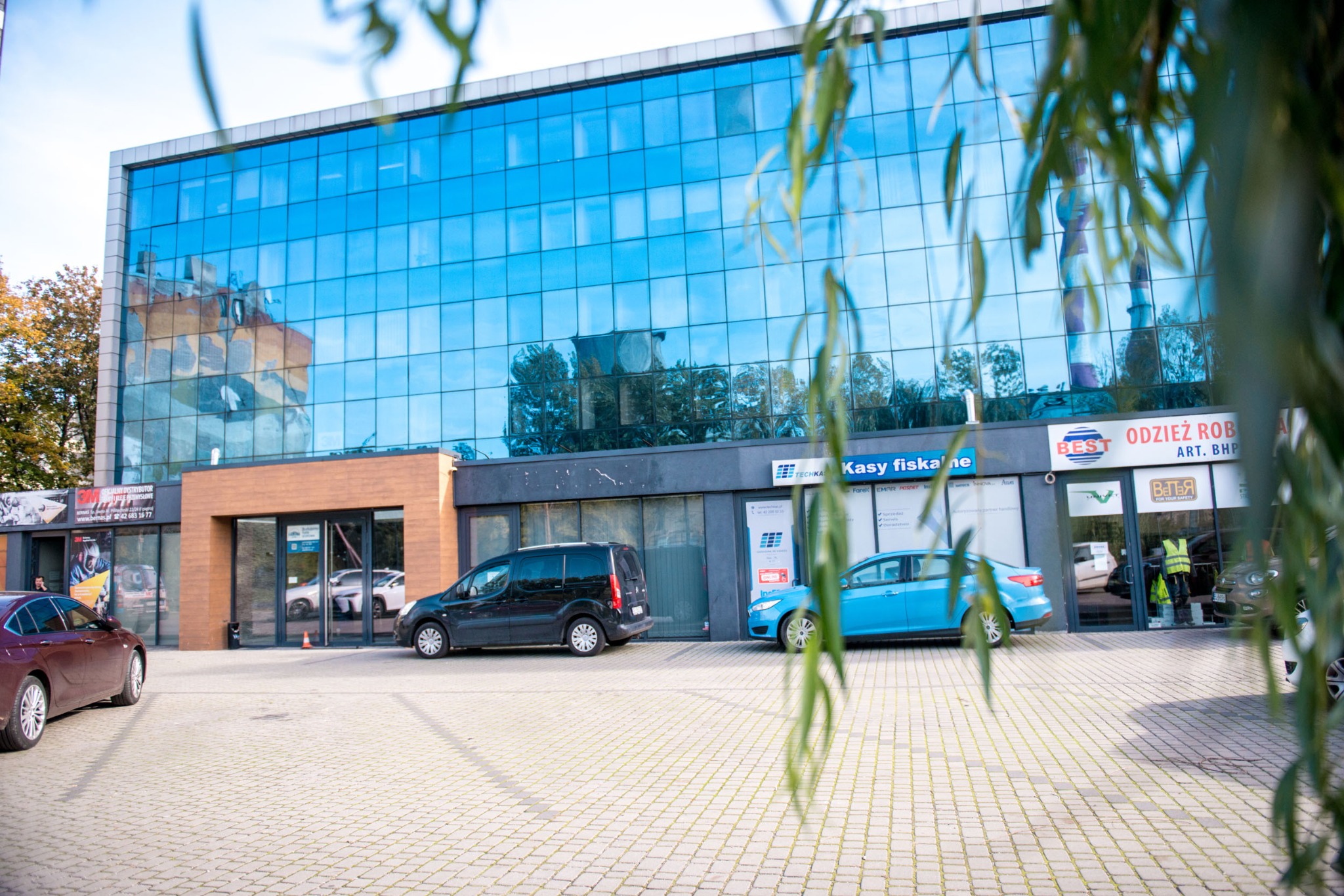 Nowoczesny budynek biurowy z przeszkloną fasadą, odbijającą niebo i otoczenie, przed którym zaparkowane są samochody, a w tle widoczne są drzewa i inne budynki.