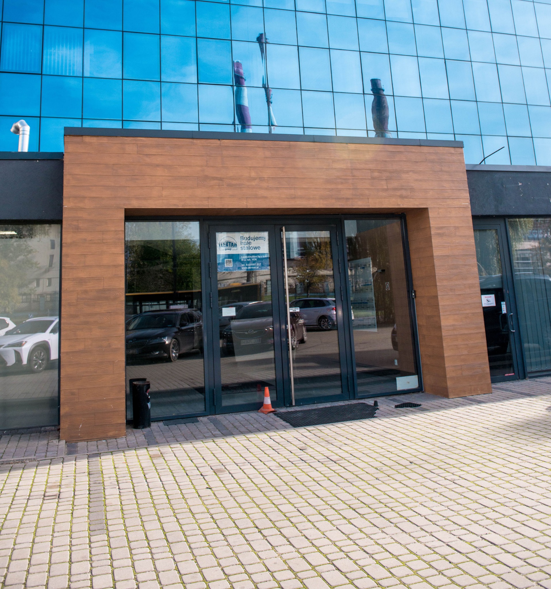 Wejście do biurowca z przeszkloną fasadą, drewnianym obramowaniem wejścia i widocznymi w szybach odbiciami samochodów. Na chodniku stoi pomarańczowy pachołek.