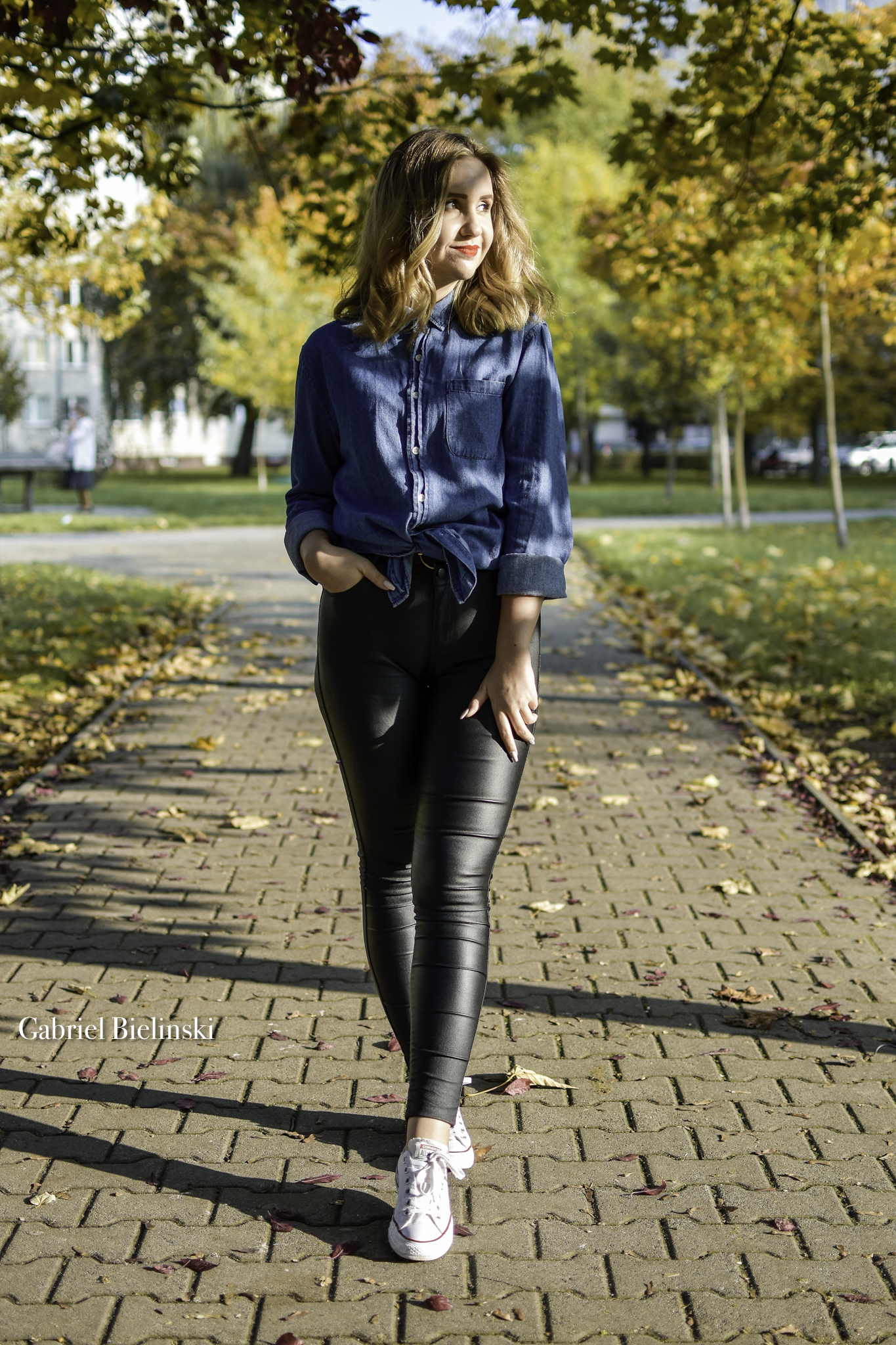 Kobieta w dżinsowej koszuli i czarnych, skórzanych spodniach idzie po chodniku w parku jesienią, z ręką w kieszeni i białych tenisówkach.