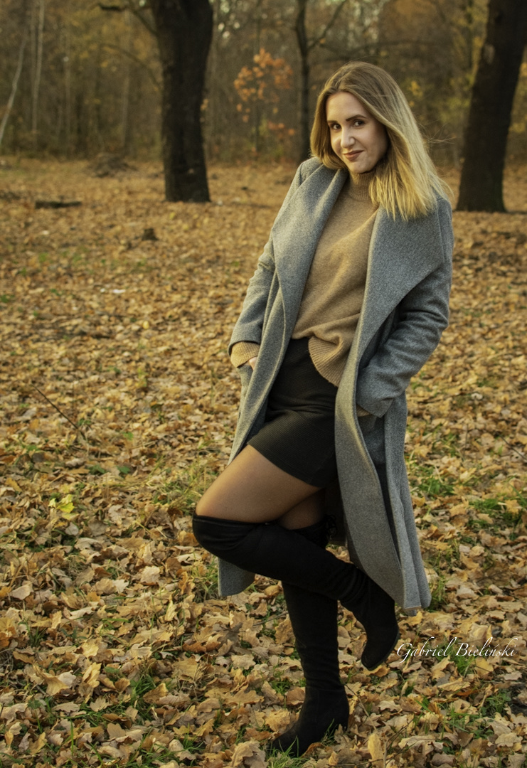 Kobieta w jesiennej stylizacji: długi szary płaszcz, beżowy sweter, czarna spódnica i kozaki za kolano, pozująca w parku pełnym opadłych liści.