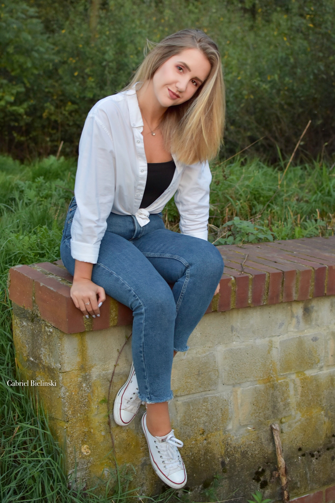 Młoda kobieta w białej koszuli i jeansach siedzi na niskim murku z cegieł, uśmiechając się w naturalnym otoczeniu z zielenią w tle.