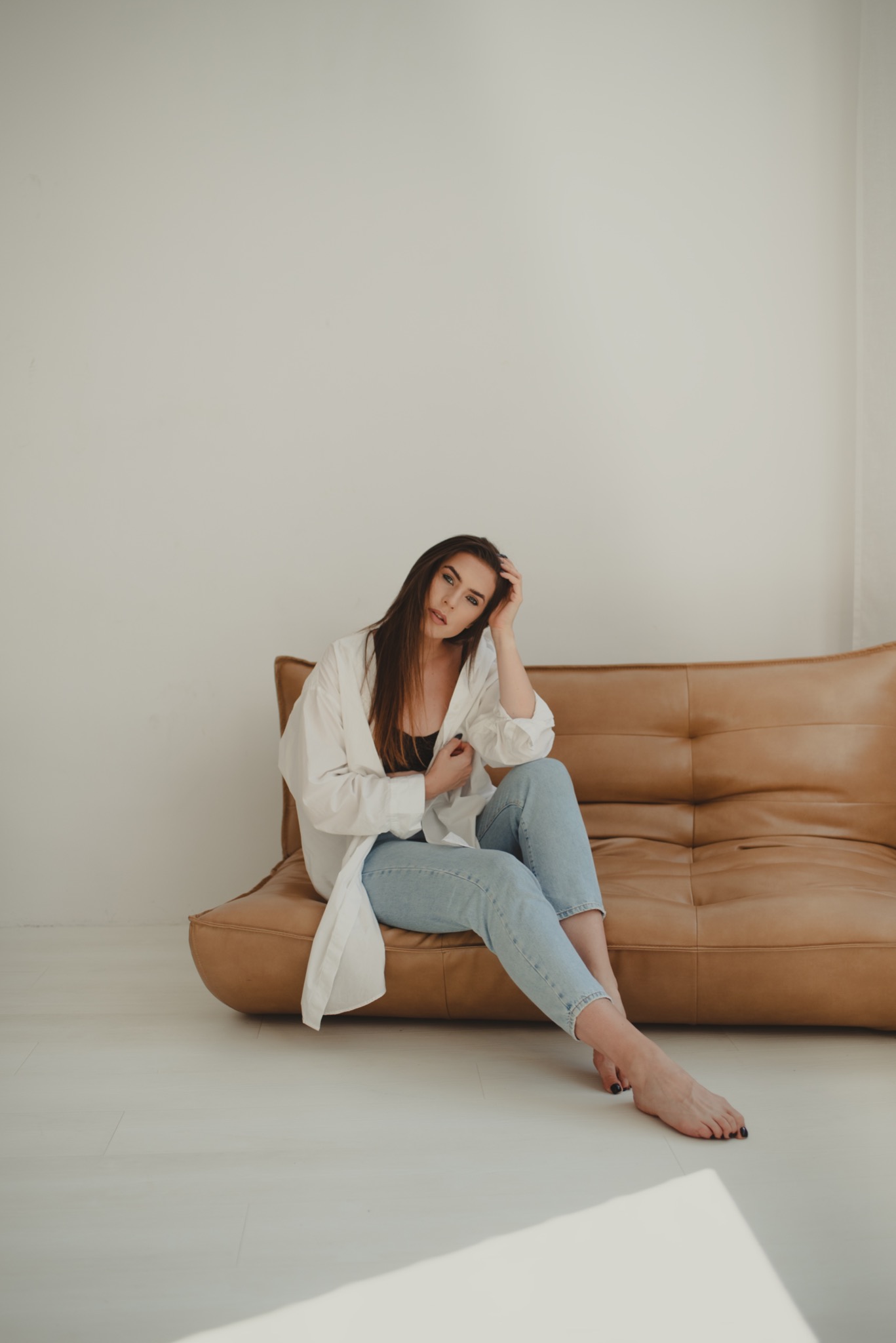 Kobieta w białej koszuli i jeansach siedzi na skórzanej sofie, opierając głowę o dłoń, z lekkim uśmiechem i spojrzeniem w obiektyw.