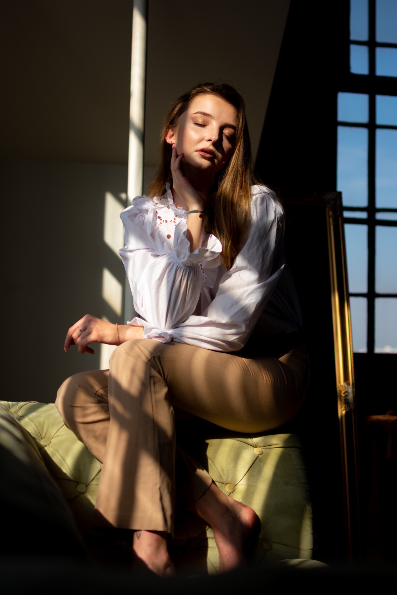 Portret kobiety w białej bluzce z koronką, siedzącej na zielonej sofie w promieniach zachodzącego słońca, z oknem w tle.