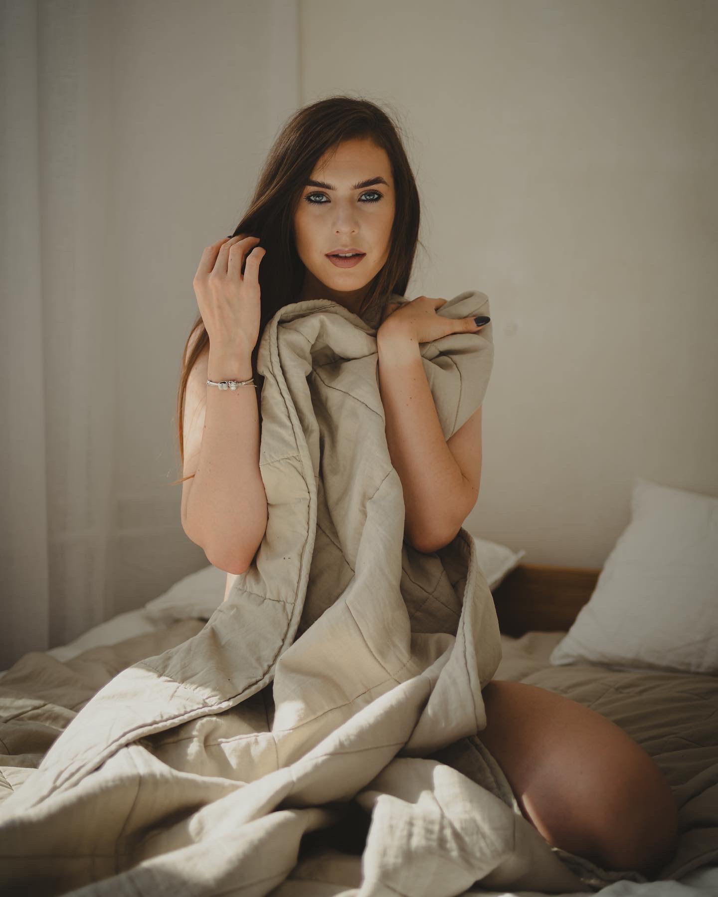 Portret kobiety z długimi brązowymi włosami i niebieskimi oczami, siedzącej na łóżku, owiniętej beżową kołdrą w naturalnym świetle.