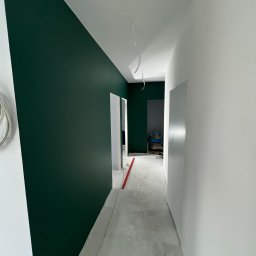 Comfort-Bud - Korytarz w trakcie malowania: jedna ściana w odcieniu butelkowej zieleni, druga biała, surowa podłoga, widoczne przewody i poziomica.
