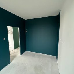 Comfort-Bud - Pokój po malowaniu ścian na ciemny turkus i biało, widoczne wystające kable elektryczne, surowa wylewka na podłodze, widok na kolejne pomieszczenie z zieloną ścianą.