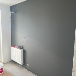 Comfort-Bud - Świeżo pomalowana szara ściana w pokoju z białym grzejnikiem, wystającymi kablami i puszkami elektrycznymi, widoczny fragment podłogi zabezpieczony kartonem.