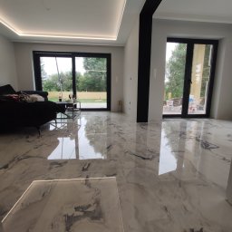 Comfort-Bud - Salon z błyszczącą podłogą imitującą marmur, czarnymi ramami okiennymi i listwami LED w suficie, z widocznym fragmentem sofy i stolika kawowego.