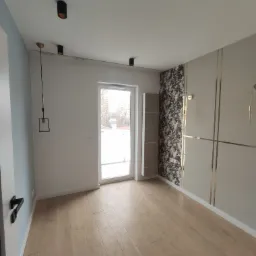 Jasne panele podłogowe w pustym pokoju z oknem balkonowym, fragmentem tapety w kwiaty i dekoracyjnymi listwami na ścianie, widoczne czarne lampy sufitowe.