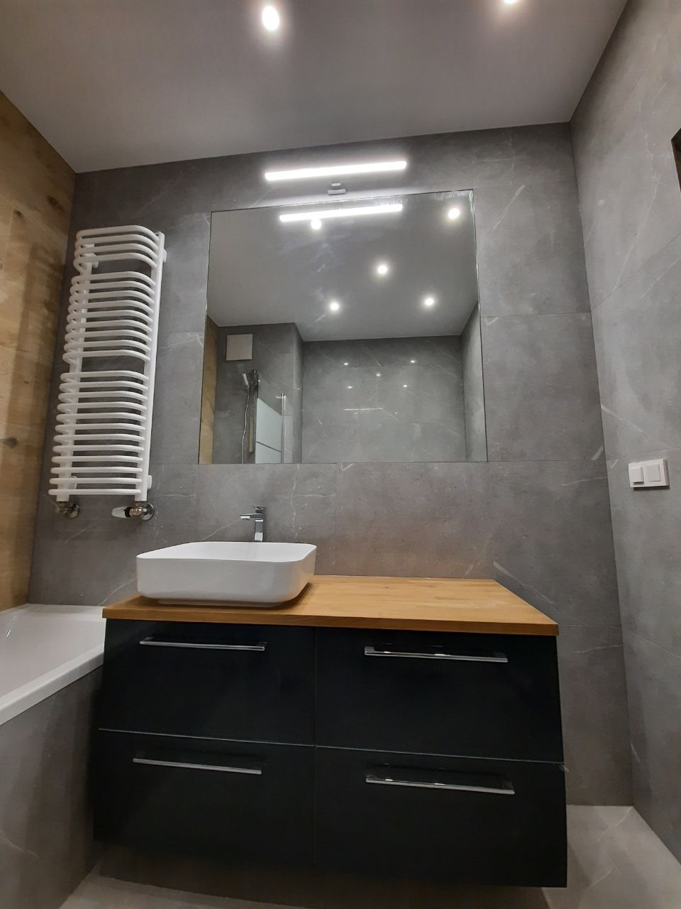 Nowoczesna łazienka z szarymi płytkami imitującymi marmur, białą umywalką nablatową na drewnianym blacie, czarną szafką pod umywalką i grzejnikiem drabinkowym.