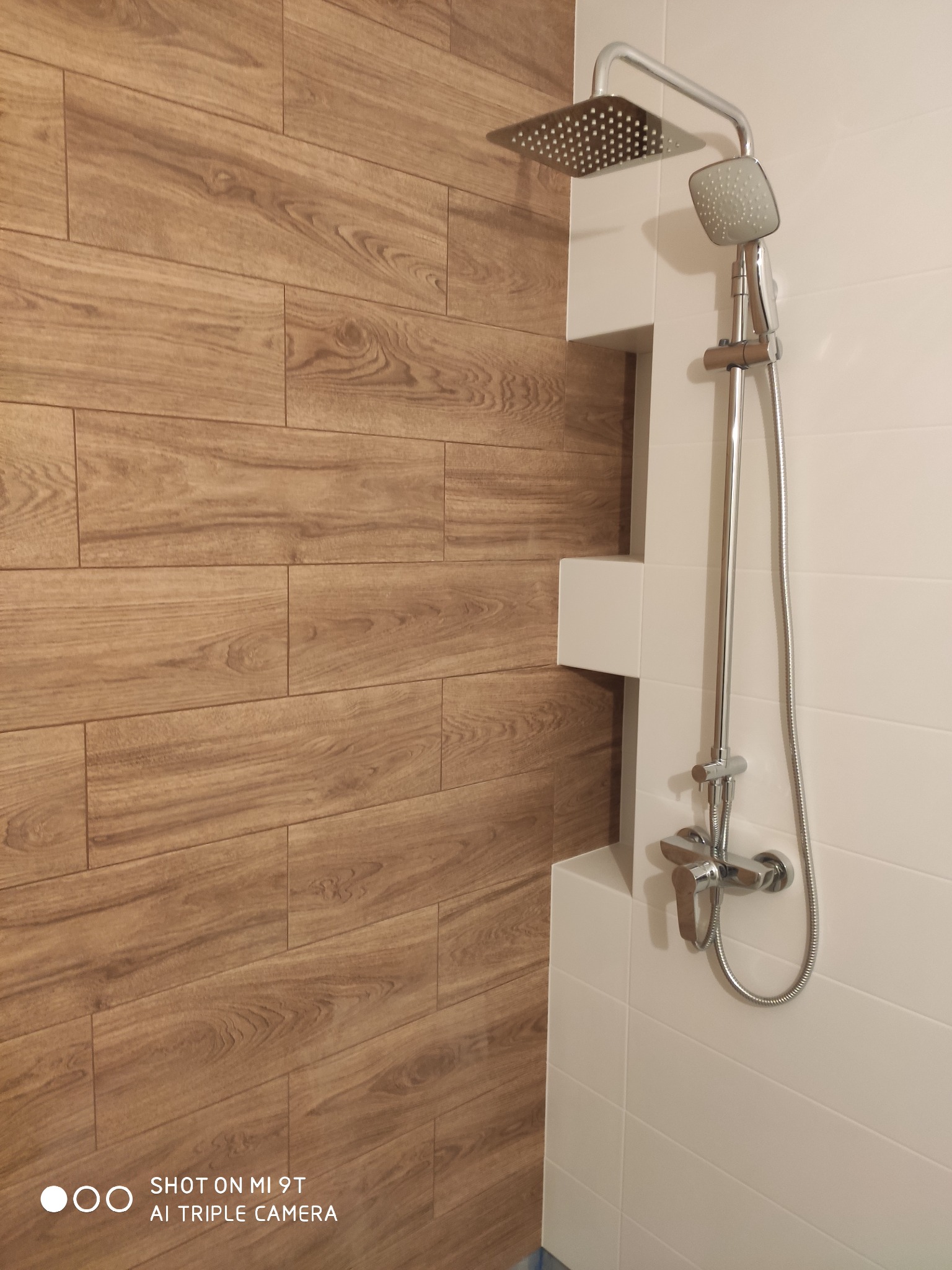 Nowoczesna kabina prysznicowa z deszczownicą i baterią, wykończona płytkami imitującymi drewno na jednej ścianie i białymi płytkami na pozostałych.