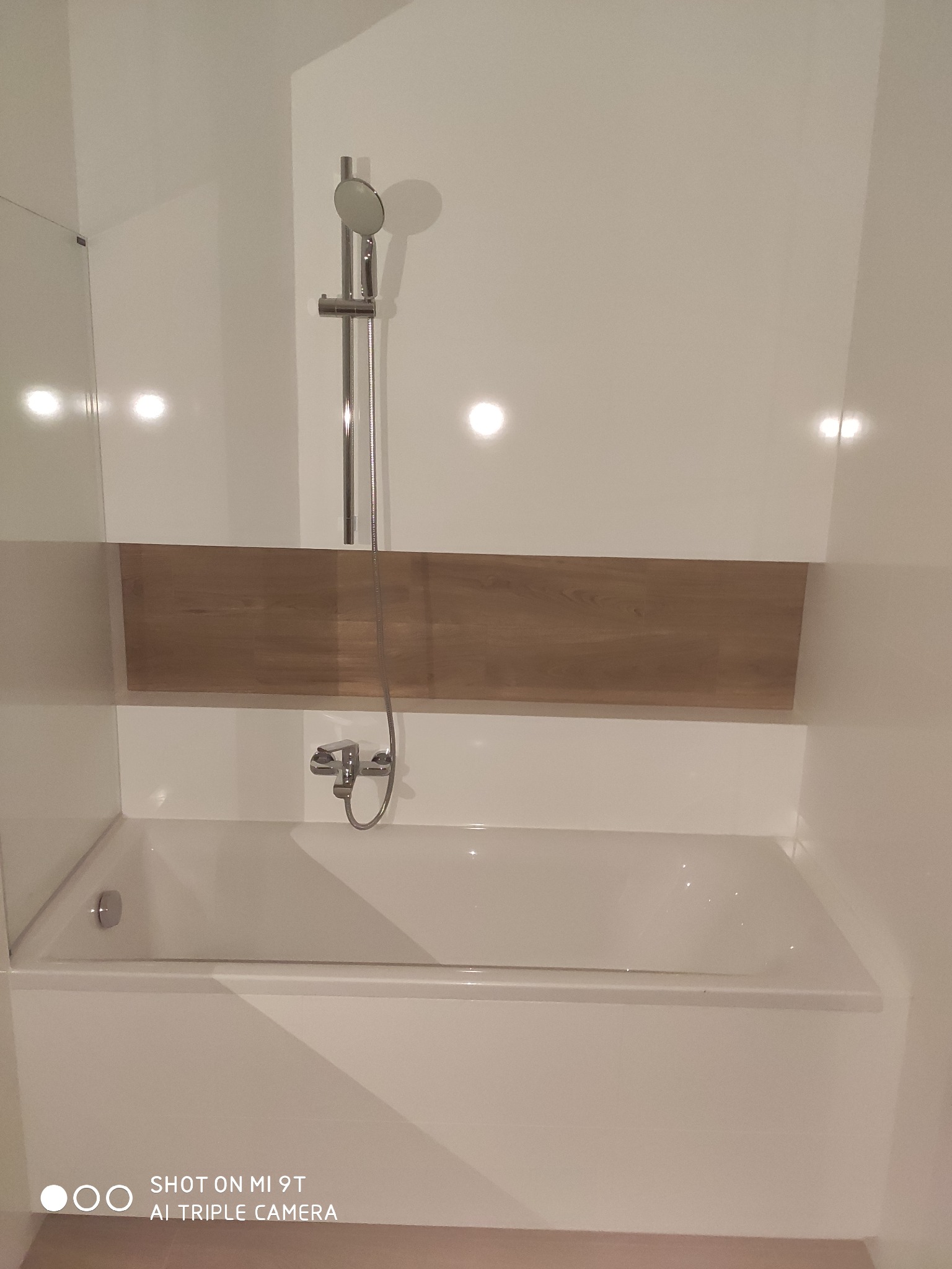 Biała wanna z drewnianym panelem dekoracyjnym i chromowaną armaturą w odnowionej łazience, widoczne odbicia światła na błyszczących powierzchniach.