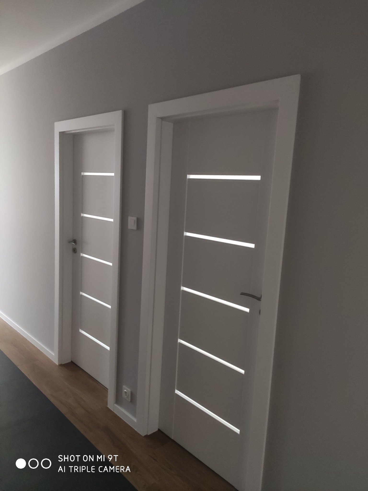 Dwa białe, nowoczesne drzwi wewnętrzne z poziomymi, podłużnymi przeszkleniami, zamontowane w szarej ścianie, widoczny fragment podłogi z panelami.